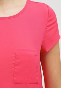 Růžová blůza s krátkými rukávy, kulatým výstřihem a malou náprsní kapsou na levé straně, vyrobená z hladké, lehké látky.