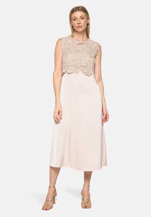 Femme debout portant une robe midi beige sans manches avec un détail en dentelle crochet sur le haut, chaussée de talons dorés à lanières sur un fond clair uni.