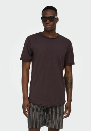 Ung mand iført sort solbriller, mørkebrun kortærmet skjorte og sorte shorts med hvide lodrette striber, stående mod en hvid baggrund.