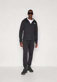 Zwarte hoodie met ritssluiting en bijpassende joggingbroek gemaakt van zachte stof. Heeft het North Face-logo op de borst en het broekspijp. Gedragen met een zonnebril.