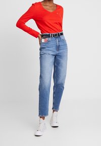 Rode lange mouwen V-hals top gecombineerd met lichtblauwe high-waisted jeans, met een zwarte riem en witte sneakers. De jeans heeft contrasterende zakdetails.