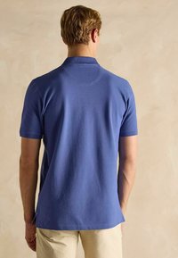 Kopā ar īsām piedurknēm izgatavota polo krekls vienkrāsainā tīra zila kokvilnā, ar apkakli, sānu šķeltiem un gludu tekstūru. Parādīts aizmugures skats.