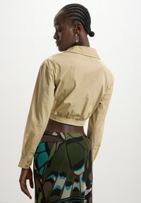 Camicia beige tagliata, con colletto e maniche lunghe, realizzata in tessuto morbido. Abbinata a una gonna con motivo che presenta tonalità verdi e blu.