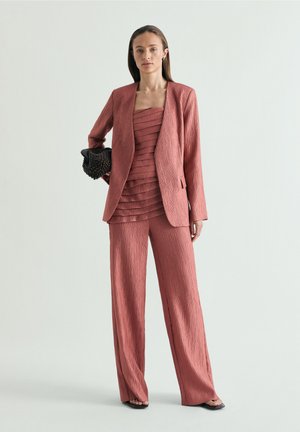 Vrouw draagt een textuurachtige poederroze pantalonpak met gelaagde top, houdt een zwarte kralenclutch vast, staande tegen een effen lichte achtergrond.