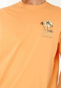 Oranges Baumwoll-T-Shirt mit kurzen Ärmeln, das ein grafisches Design von Palmen und der Sonne zeigt, und den Text "RIP CURL THE SEARCH" trägt.