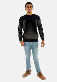 Maglione a righe navy con linee orizzontali verdi, dotato di un collo rotondo e bottoni sulle spalle; abbinato a jeans blu chiaro e stivali color sabbia.