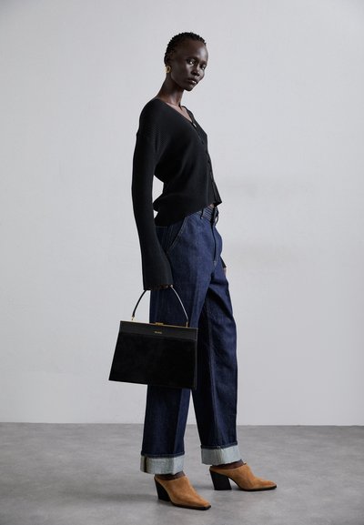 Melnā ribota džemperis, brīvi pieguļošas ziloņauduma bikses ar rullētiem dibeniem, brūni zamša zābaki ar papēžiem, tur melnu strukturētu rokassomu ar zelta akcentiem.