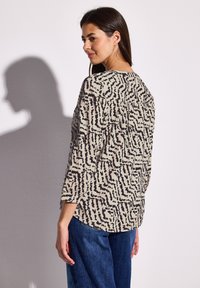 Top beige con motivo astratto nero, maniche 3/4, scollatura rotonda e vestibilità rilassata; indossato con pantaloni in denim blu.