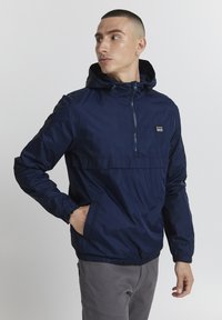 Veste coupe-vent marine avec une fermeture éclair frontale, des poignets élastiques et une capuche à cordon de serrage. Elle se distingue par un petit patch logo sur la poitrine.