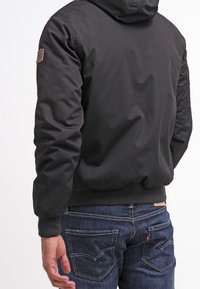 Czarna kurtka z kapturem wykonana z gładkiego materiału, z dzianinowym pasem w talii i naszywką z logo na lewym rękawie. Noszona z niebieskimi dżinsami.