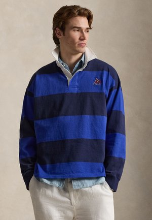 Młody mężczyzna z czerwonymi włosami, ubrany w niebieską i granatową koszulkę rugby w paski na jasnoniebieską koszulę na guziki i beżowe spodnie, patrzący w prawo.