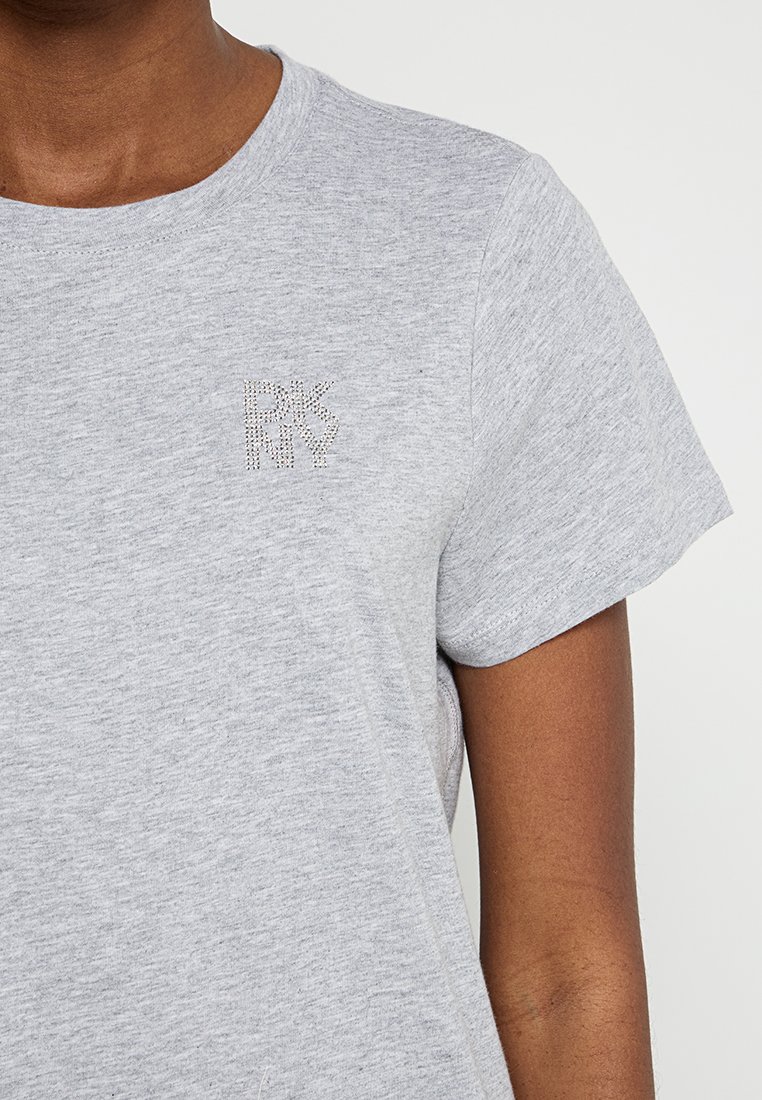 Osoba ubrana w jasnoszary T-shirt z krótkim rękawem, z małym dekoracyjnym logo "R N K Y" wykonanym z kryształków na piersi.