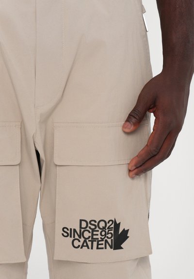 Pantalons cargo beiges avec deux grandes poches, arborant le texte noir "DSQ2 SINCE 95 CATEN" et des détails accentués. Texture lisse, tissu léger.