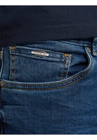 Blå jeans i denim med gyllene sömmar och en liten silveretikett på fickan märkt "PETROLIND". Slät textur och klassisk design.