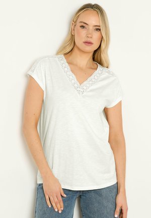 Blonde Frau trägt ein weißes V-Ausschnitt-Kurzarmoberteil mit Spitzenbesatz und blaue Jeans, steht vor einem hellen Hintergrund.