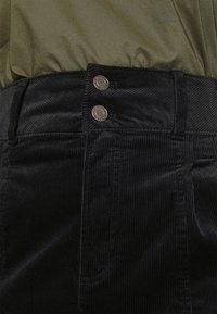 Pantalon en velours côtelé noir taille haute avec deux boutons métalliques et passants de ceinture visibles, associé à un haut vert olive.