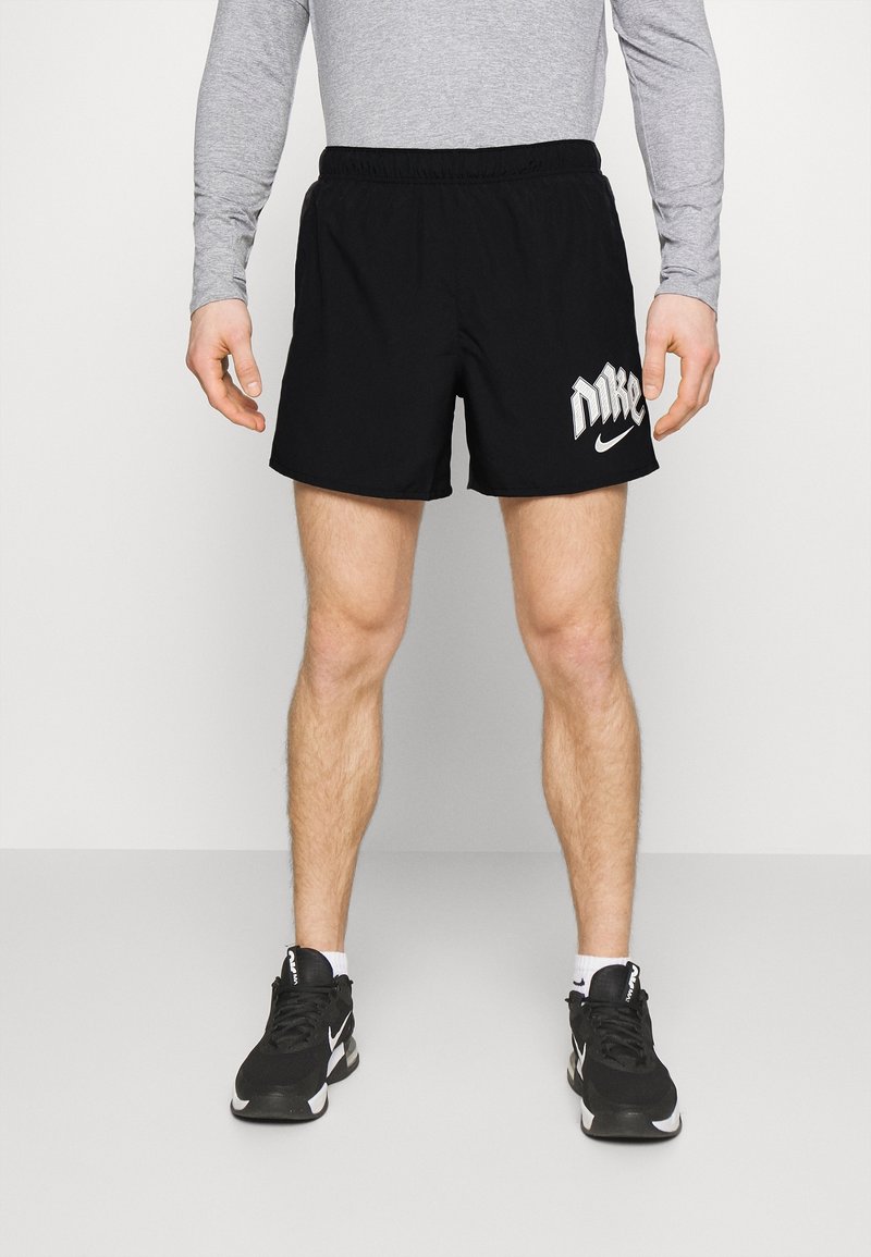 Hombre vestido con pantalones cortos negros de Nike, camiseta de manga larga gris, calcetines blancos y zapatillas negras de Nike, de pie contra un fondo liso.