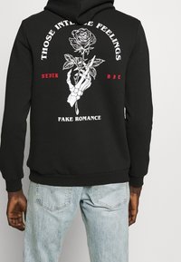 Svart hoodie med en grafisk bild av en skelett-hand som håller en ros. Texten inkluderar "THOSE INTENSE FEELINGS" och "FAKE ROMANCE" i rött och vitt.