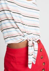 Biały prążkowany top z wielokolorowymi paskami w odcieniach różu, niebieskiego i zielonego, związany w talii, w połączeniu z czerwoną spódnicą z detalami w postaci guzików.