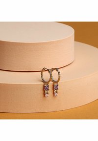 Guld hoop øreringe har et snoet design med farverige firkantede ædelstene i lilla, grøn og pink, præsenteret på strukturerede ferskenfarvede pedestaler.