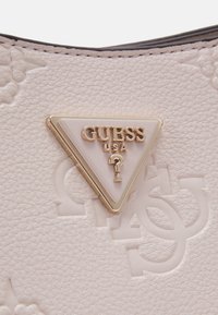 Borsa a tracolla in pelle beige con texture cremosa e motivi embossati, caratterizzata da una placca triangolare in oro e bianco con il logo "GUESS USA".