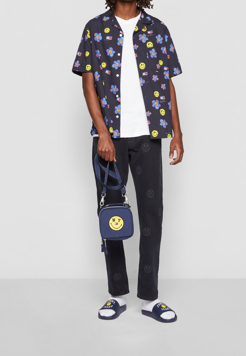 Homme portant une chemise noire à fleurs et smileys, un t-shirt blanc, un pantalon noir avec des imprimés smiley, tenant un sac bleu marine avec un smiley, des sandales bleu marine avec des chaussettes.