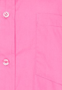 Camisa rosa clara de botão com um bolso no lado esquerdo do peito, apresentando botões redondos rosa e uma textura de tecido suave.