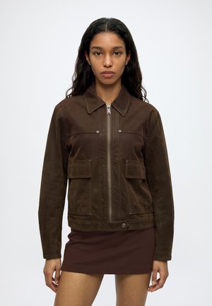 Jeune femme aux longs cheveux foncés portant une veste en daim marron zippée et une jupe marron assortie, debout devant un fond blanc uni.