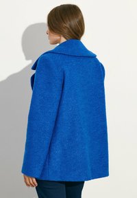 Nainen, jolla on ruskea hius ja kirkkaan sininen teksturoitu takki, seisoo kyljellään yksinkertaista vaaleaa taustaa vasten.