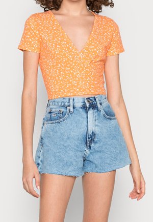 Femme portant un top court orange à manches courtes avec de petites fleurs blanches et un short en jean bleu clair taille haute.