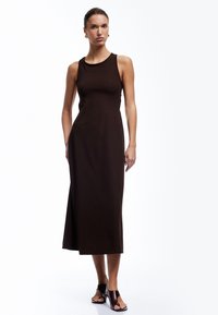 Vestido marrón, sin mangas, de largo hasta la rodilla, con una silueta ajustada y una abertura lateral. Combinado con sandalias de slides negras y burdeos.