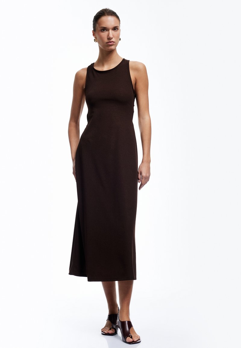 Vestido marrón, sin mangas, de largo hasta la rodilla, con una silueta ajustada y una abertura lateral. Combinado con sandalias de slides negras y burdeos.
