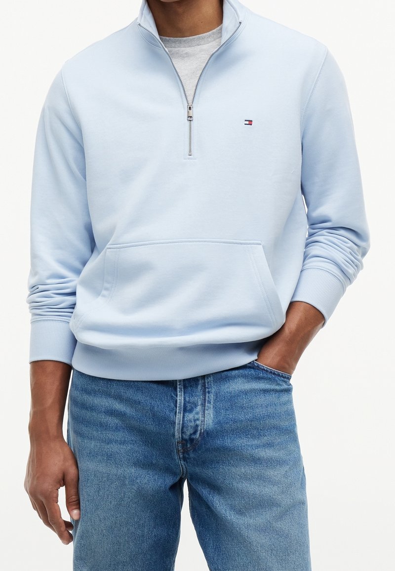Personne portant un sweat-shirt bleu clair avec fermeture éclair au quart et petit logo sur la poitrine, ainsi qu'un jean bleu clair, une main dans la poche, sur fond blanc.