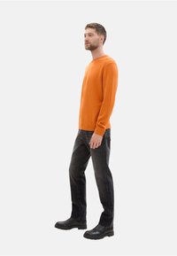 Suéter naranja con puños acanalados, combinado con jeans oscuros y botas de tobillo negras. El suéter tiene un cuello redondo y un corte clásico.