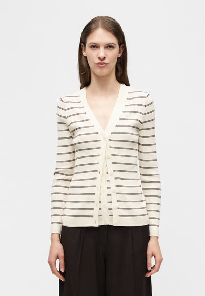 Γυναίκα που φοράει μπεζ ζακέτα με λεπτές σκούρες οριζόντιες ρίγες και μαύρο παντελόνι, στέκεται μπροστά σε απλό ανοιχτόχρωμο φόντο.