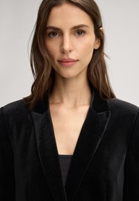 Donna con pelle chiara e capelli castani che indossa un blazer di velluto nero e un top nero, che guarda dritto davanti a sé su uno sfondo chiaro e uniforme.