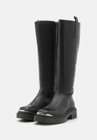 Botas negras de cuero lisas hasta la rodilla con cierre lateral, que cuentan con una suela gruesa y un acento metálico en la puntera. Parte superior texturizada.