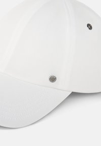 White baseball cap with textured fabric, curved visor, stitched detailing, and a metallic accent near the top.