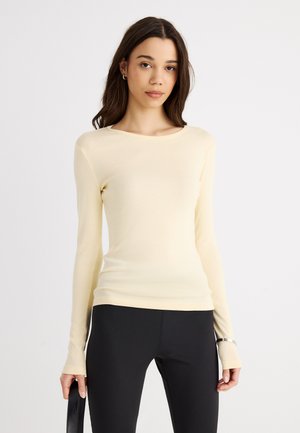 Mlada ženska z dolgimi temnimi lasmi nosi kremno dolgo majico z dolgimi rokavi in črne hlače, v levi roki drži črno naramnico.