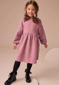 Rosa langærmet kjole med tekstureret blomstermønster, indsnævret talje og blødt stof. Kombineret med sorte knæhøje støvler og mørke strømpebukser.