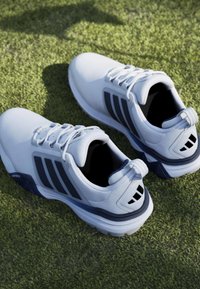 Ζευγάρι λευκά αθλητικά παπούτσια με σκούρες ρίγες τοποθετημένα σε πράσινο χλοοτάπητα, που δείχνουν θέες από επάνω και από το πλάι.