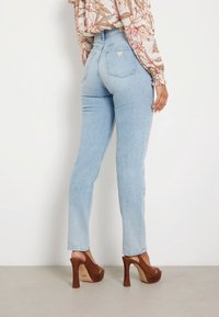 Mujer vistiendo jeans de tiro alto en azul claro con bolsillos traseros y sandalias de plataforma de tacón alto marrón, de pie contra una pared blanca.