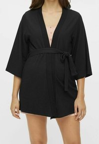 Svart texturerad robe med breda ärmar, ett bälte i midjan och V-ringning, lager på lager över ett ljust rosa plagg med knappar.