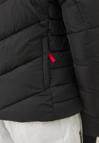 Μαύρο μπουφάν puffer με διαγώνια πηγή. Διαθέτει μια αντίθετη κόκκινη υφασμάτινη ταινία στο πλάι. Το υλικό φαίνεται μαλακό και μονωμένο.