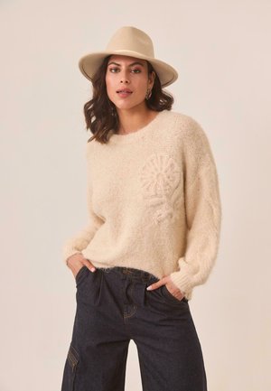 Suéter de punto beige con un diseño de flores texturizadas en el pecho, combinado con pantalones de mezclilla oscuros. El sombrero es de fieltro de color claro con un ala ancha.
