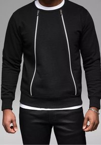 Sudadera negra con detalles de cremallera blanca, cuello redondo y puños acanalados. Textura suave y diseño ajustado, llevada sobre una camisa blanca.
