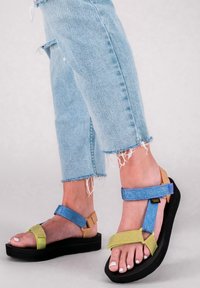 Des pieds portant des sandales Teva bleues et jaunes avec des semelles noires, associées à un jean déchiré de couleur bleu clair.