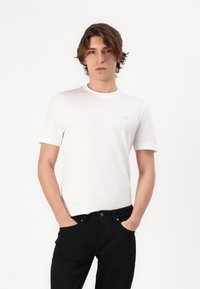 T-shirt en coton blanc à manches courtes, col rond, avec un petit logo bleu clair sur le côté gauche, associé à un jean noir.