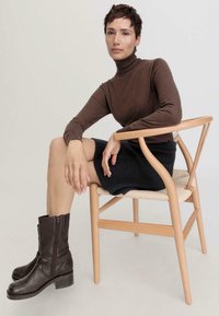 Pull en cachemire marron ajusté, jupe noire côtelée, bottines en cuir marron à lacets, assise sur une chaise en bois clair, fond minimaliste.