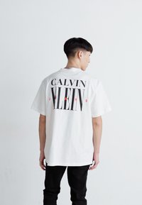 Valge puuvillane T-särk, mille seljal on julge must tekst "CALVIN KLEIN JEANS" punaste aktsentidega. Lõdva istuvuse disain.